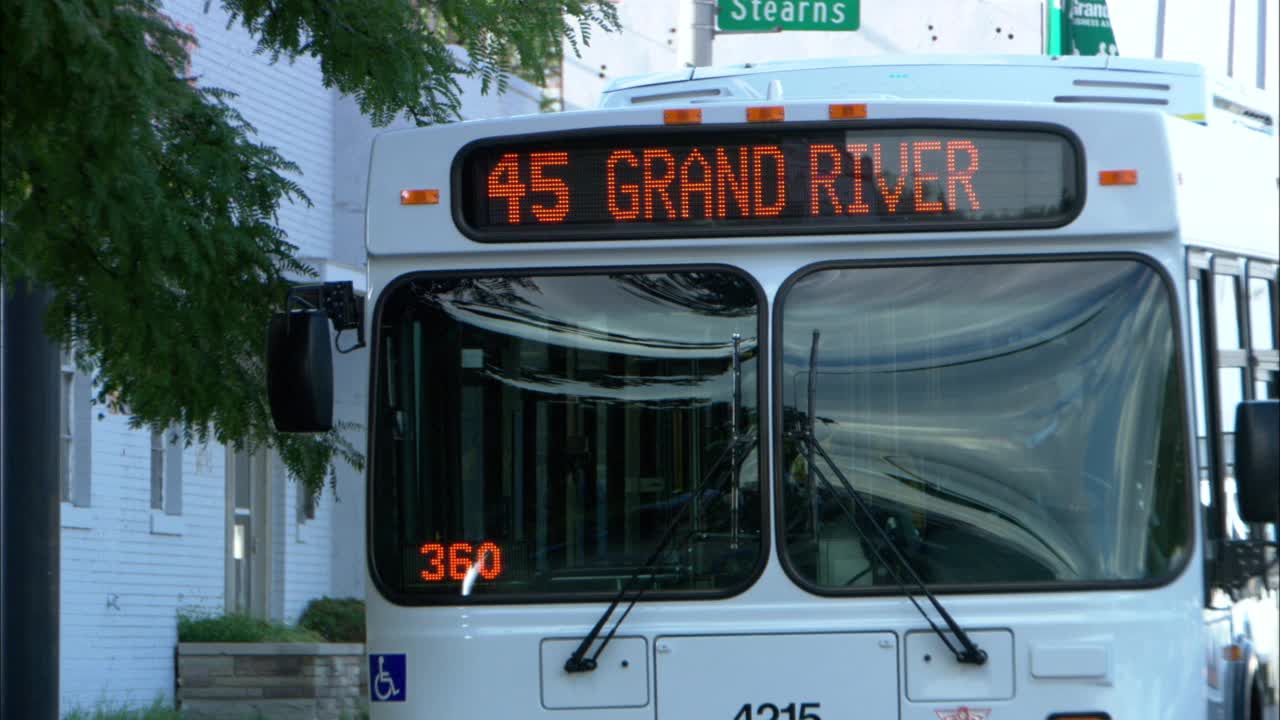 中等角度的城市公共汽车停在公共汽车站或街角。巴士上的路标在“45 7英里”和“45大河”之间交替。底特律。总线是白色的。背景是白砖建筑。风吹过左边的树视频素材