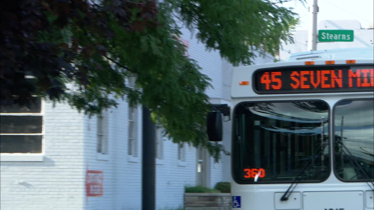 放大一辆停在公交车站或街角的城市巴士。路标在"45 7英里"和"45大河"之间交替。"底特律。公共汽车上方可见写有“斯特恩斯”的路标。背景是白砖建筑。风从树上吹过视频下载
