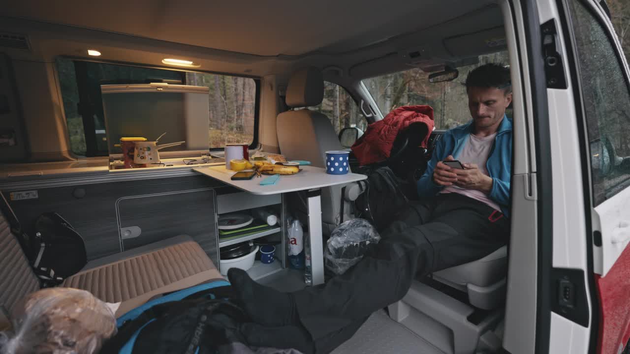 一名棕色短发的男子在他的面包车里一边用手机一边伸开双腿视频下载