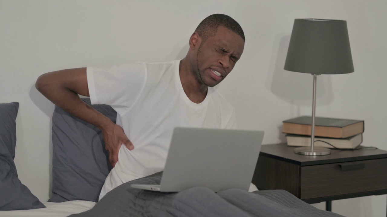 一名背部疼痛的非洲男子在床上用笔记本电脑工作视频素材