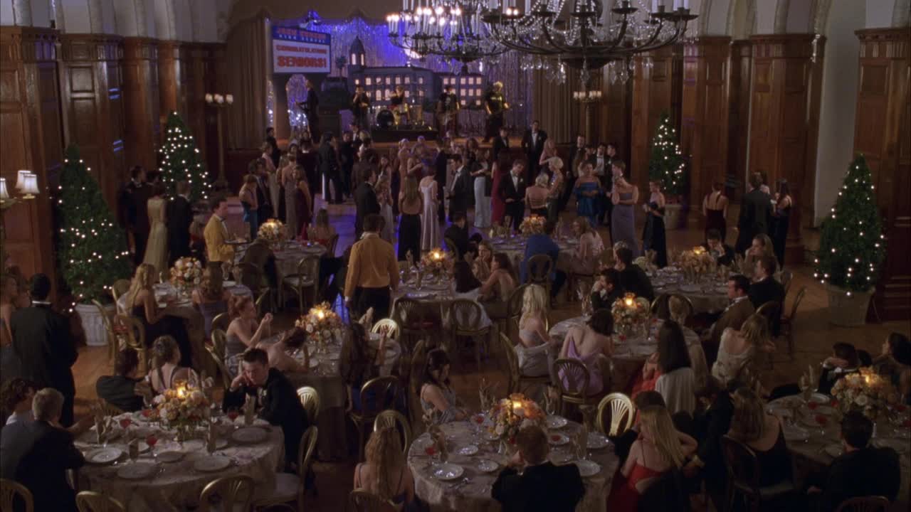 宴会用餐区角度适中。看到穿着黑色西装和连衣裙的人们围坐在摆着蜡烛和鲜花的桌子旁。看背景乐队在舞台上演奏。见吊灯顶架。看见房间两边的亮着灯的松树。SE视频素材