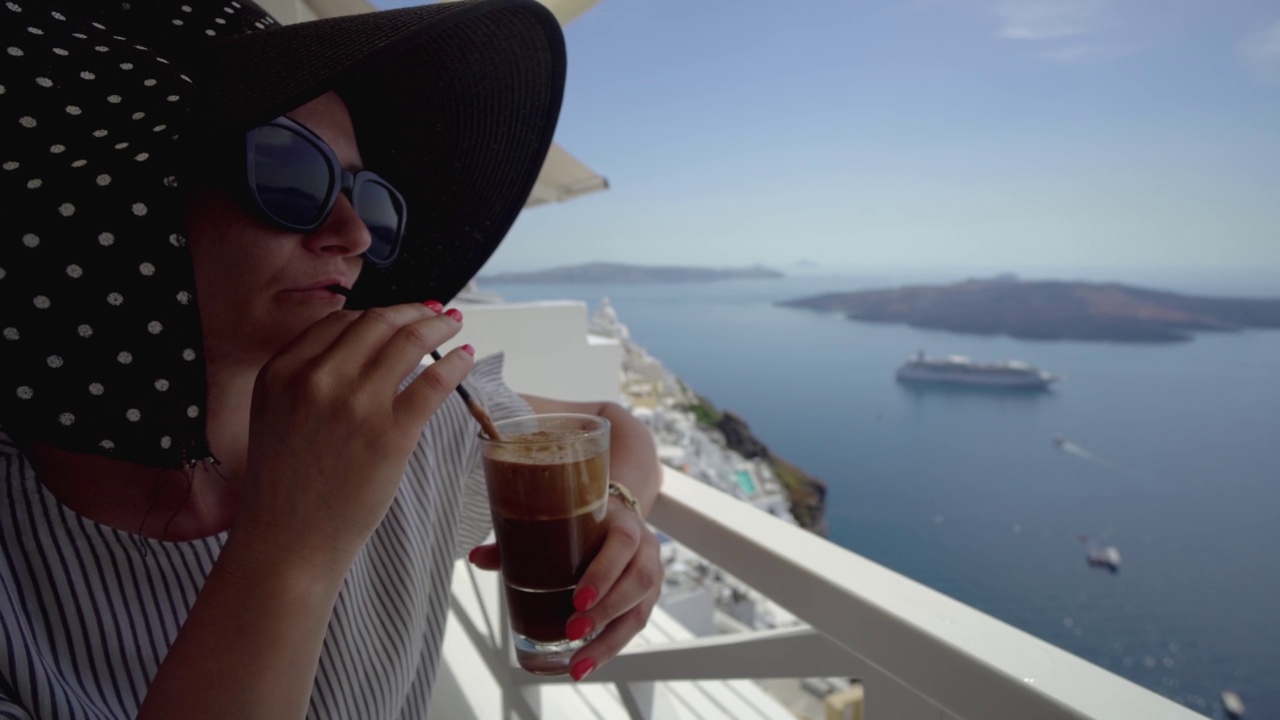希腊圣托里尼度假时喝星冰乐咖啡的女人。从餐厅阳台俯瞰火山口和火山的美景视频下载