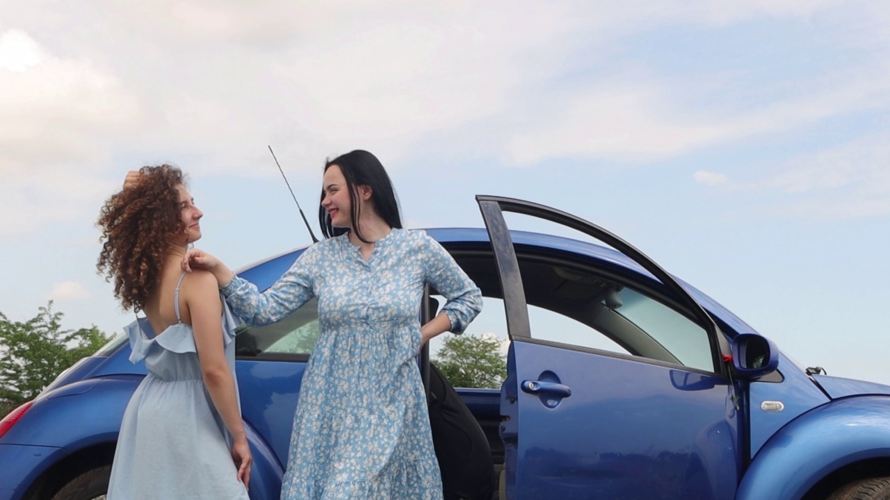 两个年轻漂亮的女孩站在田野里一辆蓝色的汽车旁边。两个女孩开着一辆蓝色的车。4 k的决议视频下载