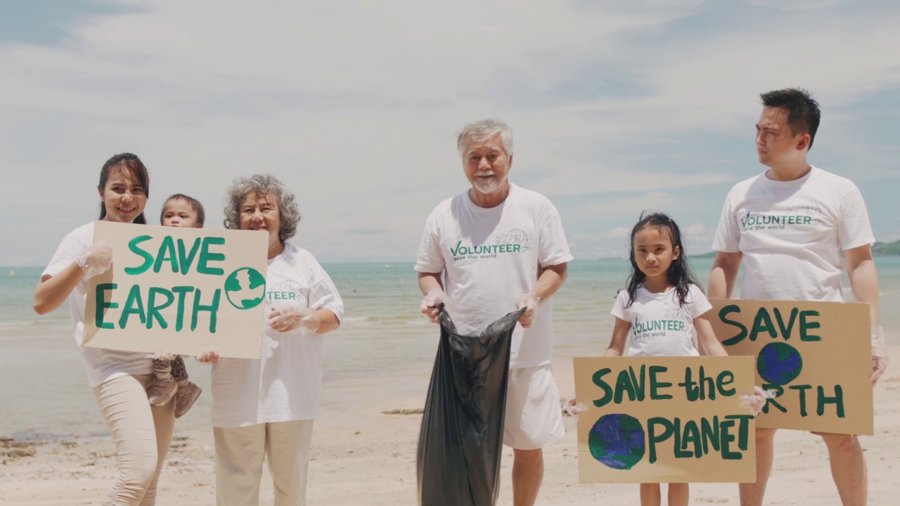 幸福的亚洲大家庭穿着志愿者衬衫，在沙滩上培养孩子们学习保护自然。一个大家庭拿着纸板，为保护地球免受环境问题的影响而奔走。视频下载