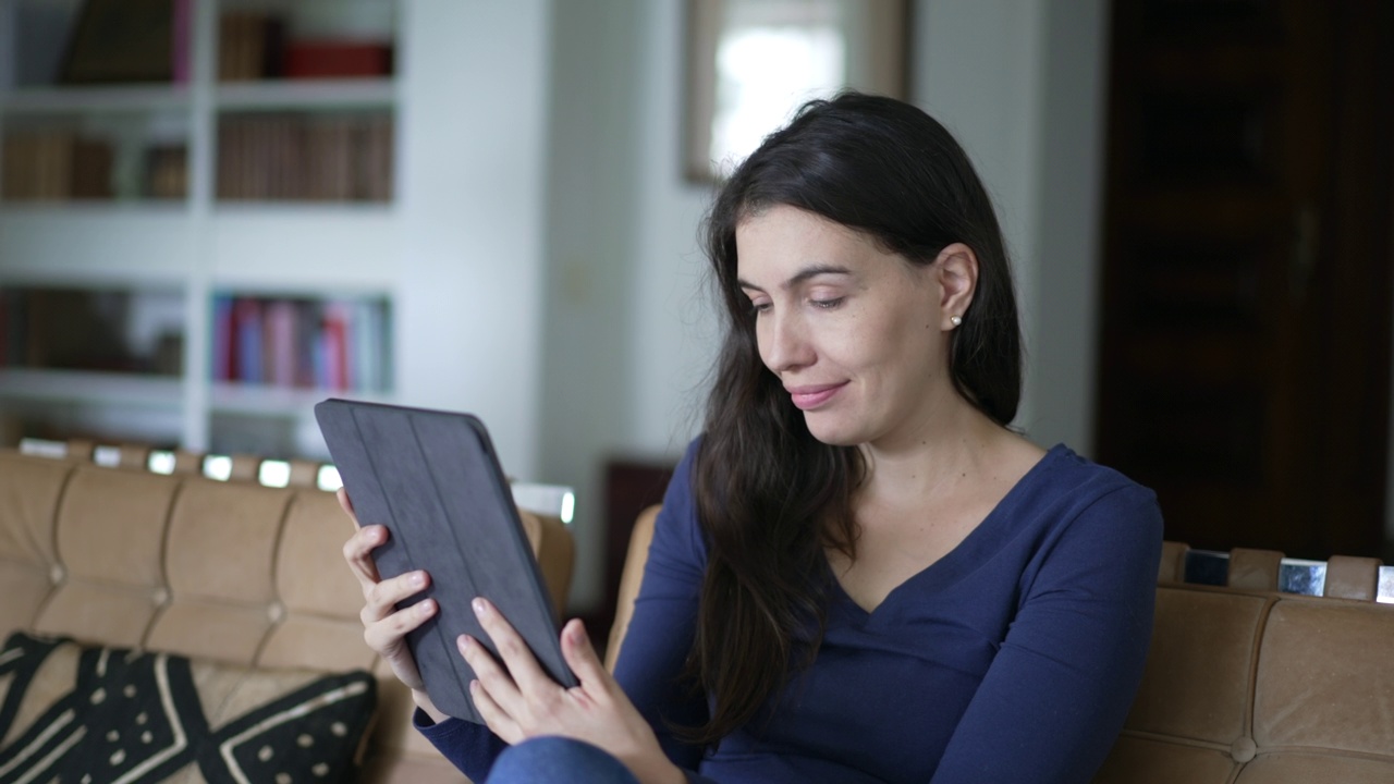 幸福的女人在家里客厅拿着平板电脑浏览互联网。使用现代数码设备的人对着媒体内容微笑视频下载