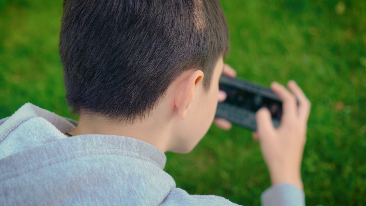 背后看男孩对手机游戏的依赖。孩子不能扔平板电脑和在户外玩视频素材