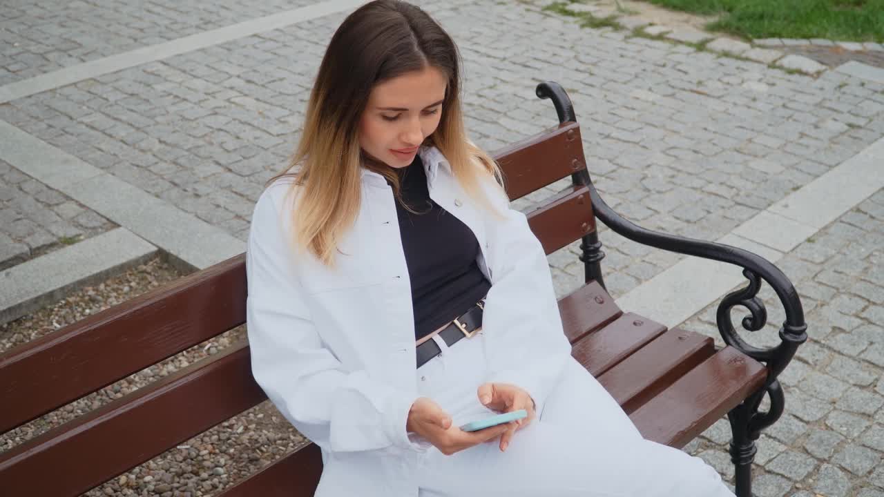 快乐的潮女人的肖像在户外城市长凳上用手机滚动社交媒体视频素材
