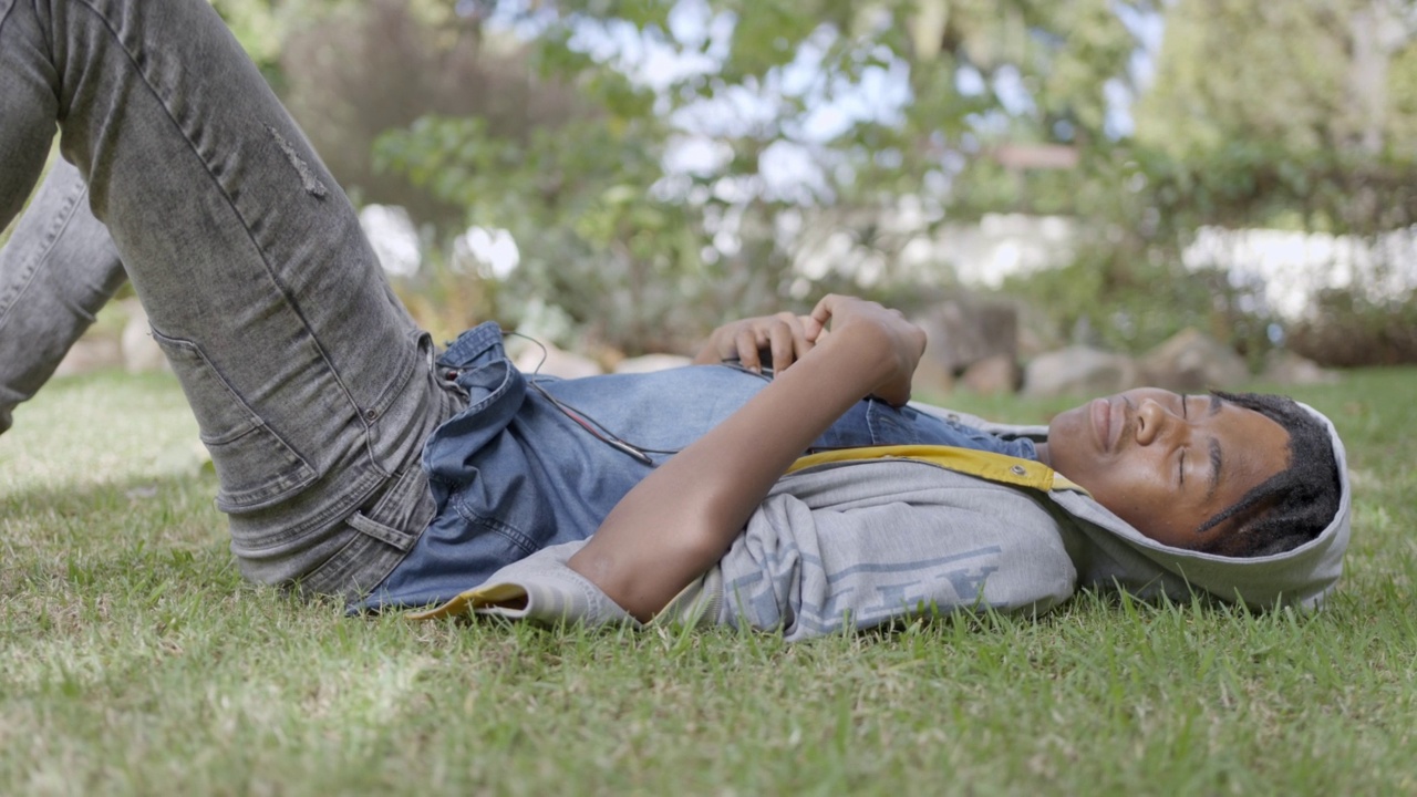 少年躺在公园里，戴着耳机听智能手机的歌单视频素材