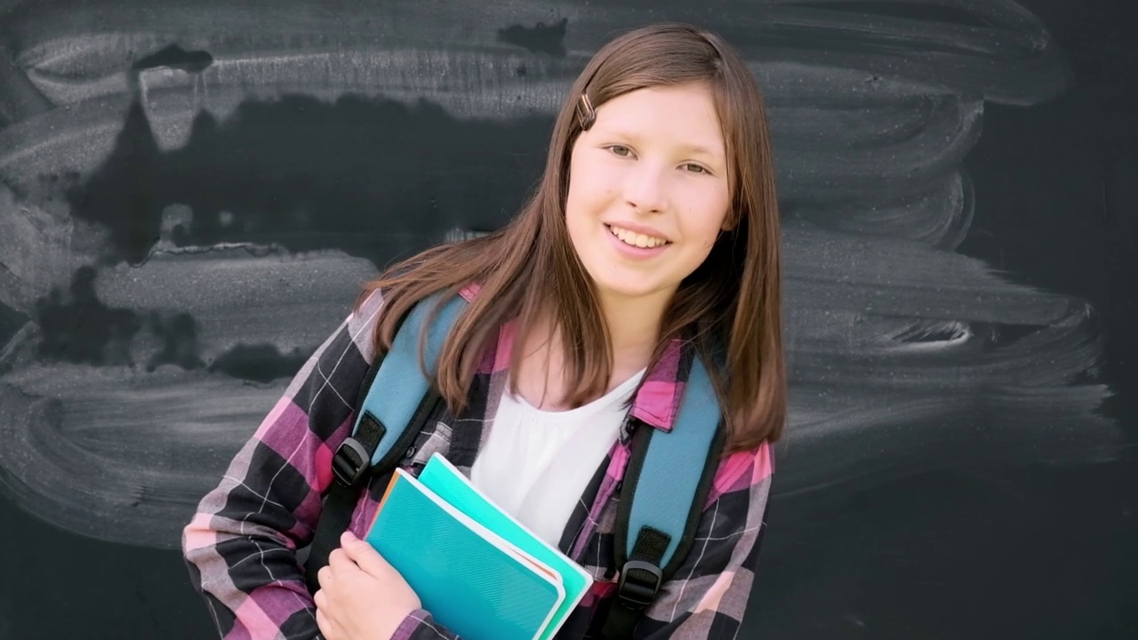 十几岁的学生女孩在衬衫，眼镜与蓝色的背包和教科书在黑色黑板的背景。教育、学校和人民。快乐快乐的少女在黑板旁跳舞，跳跃视频下载