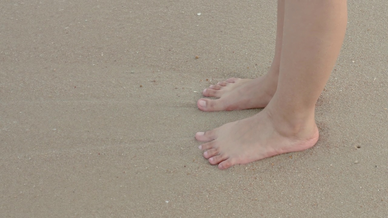 4K，一名亚洲女性站在海滩沙滩上的特写，这是一个海滨景点，与此同时，海水拍打着她的脚。感受海风吹来的凉爽视频下载