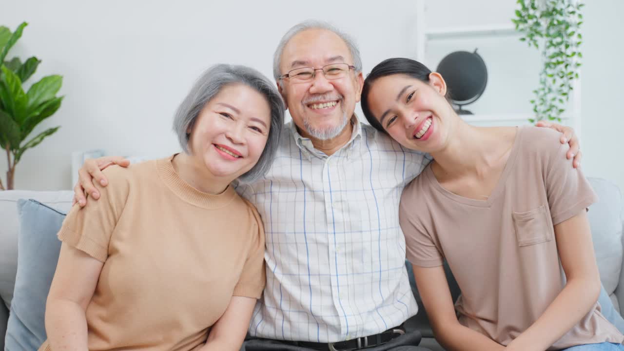 亚洲老年夫妇与年幼的女儿坐在房子里的肖像。迷人美丽的孙女探望年迈成熟的祖父母，然后微笑着，在家里的客厅一起看镜头视频素材