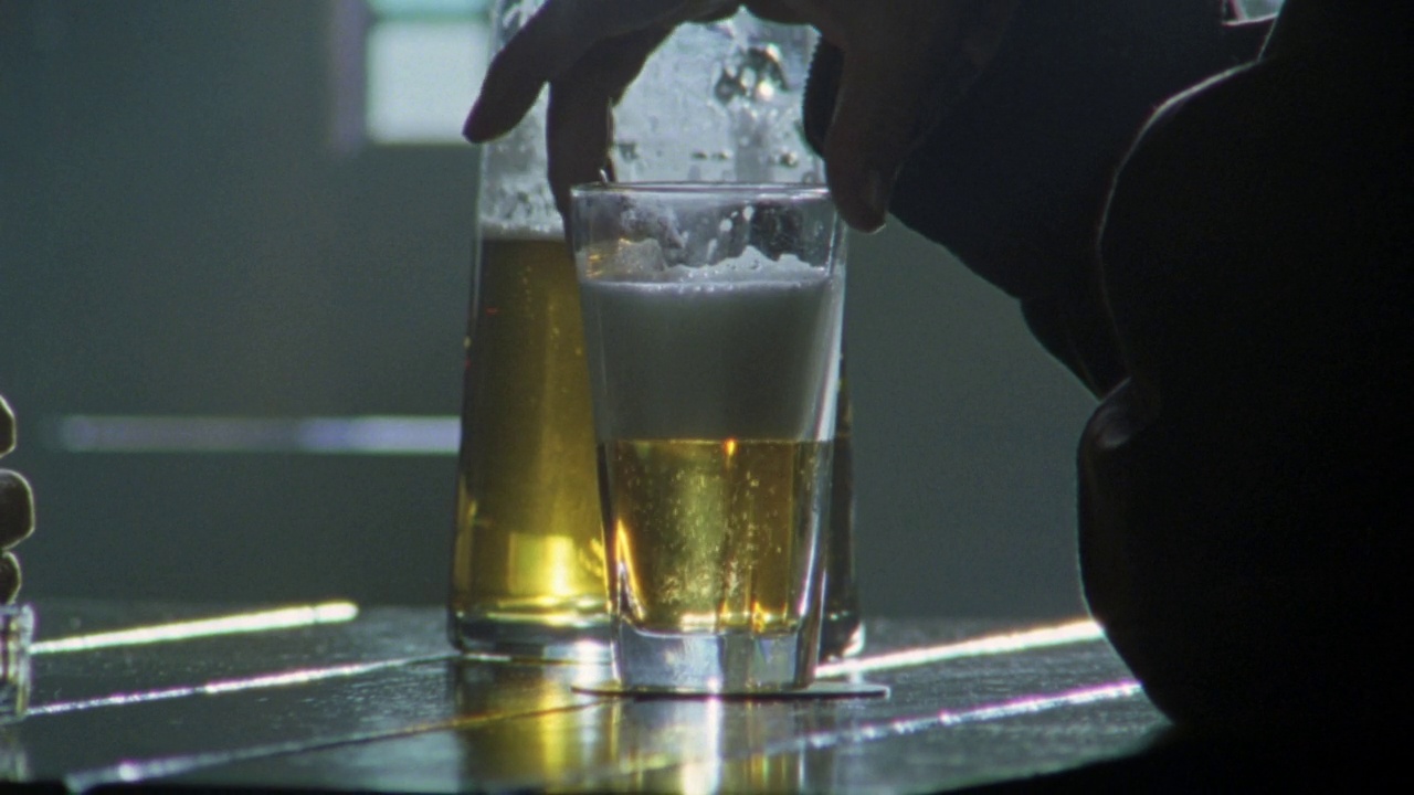 酒吧或酒馆桌子的近角。两个男人坐在桌子对面，双手从大壶往玻璃杯里倒啤酒，喝着酒。的谈话。Negative有点脏视频素材