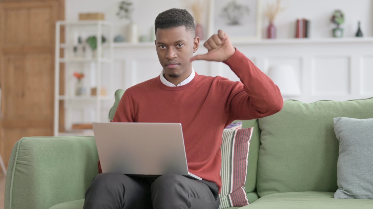 一名非洲男子在沙发上使用笔记本电脑时表现出大拇指向下的手势视频素材