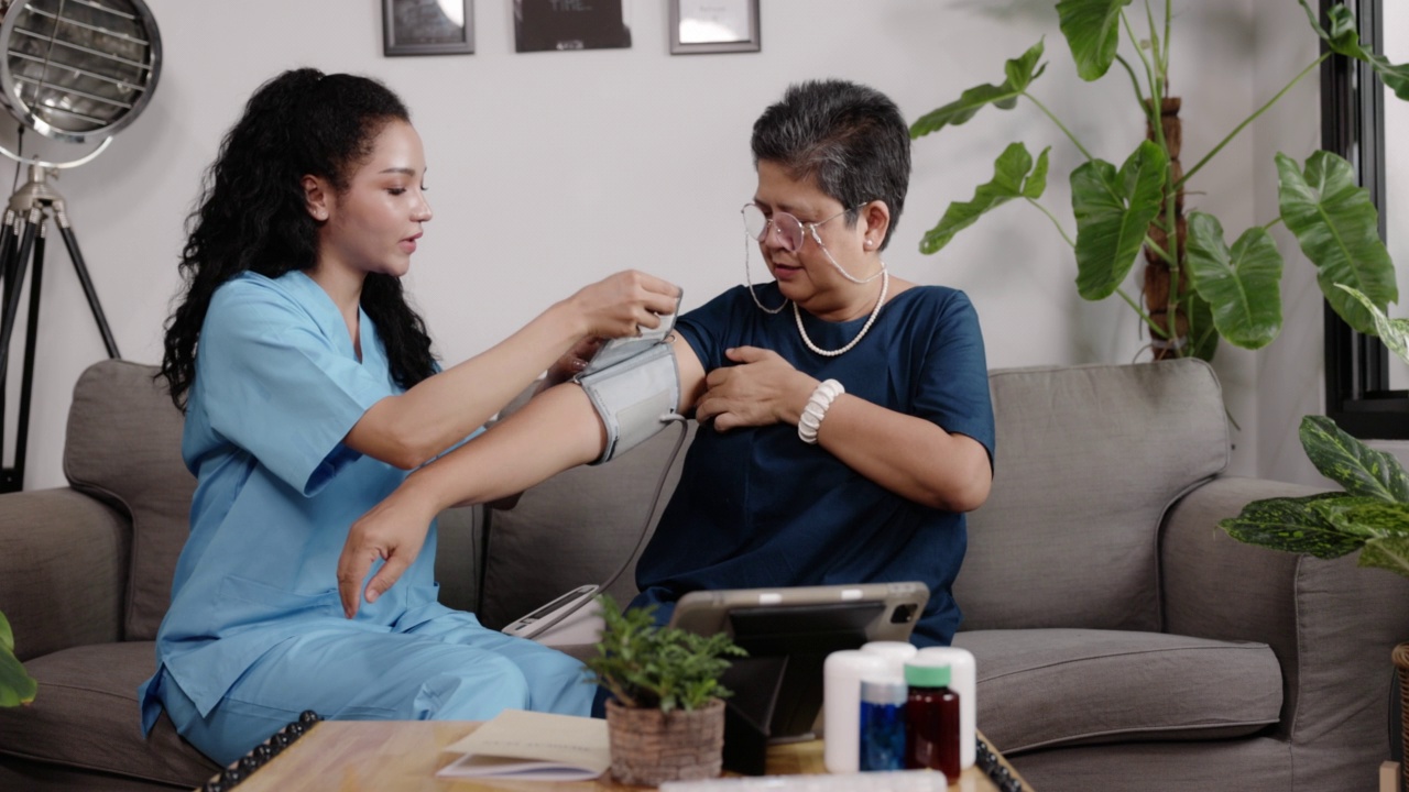 在家里的客厅里，护士在一位亚洲老人身上使用血压计。在家提供医疗服务。照顾老年病人。个人护理业务。养老院视频素材