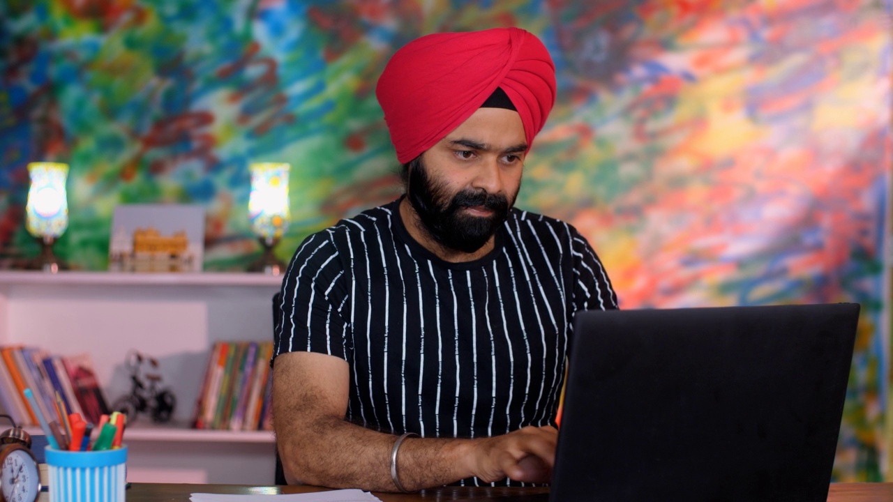 一个穿着休闲服装、戴着红色头巾的聪明的印度人在他的笔记本电脑上工作——一种数字设备，办公室工作视频素材