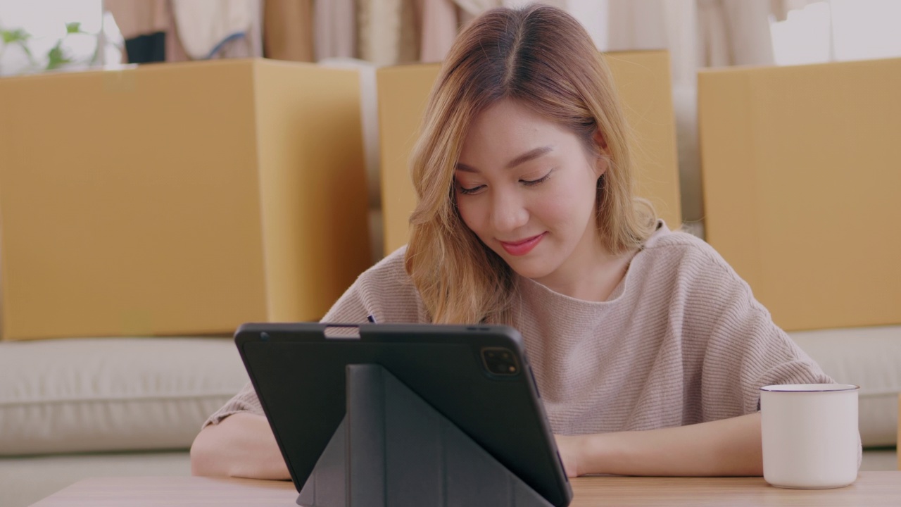 亚洲年轻女性通过Livestream在网上卖衣服，充满乐趣和快乐。度假时坐在地板上用智能手机在家直播的年轻女子。视频下载