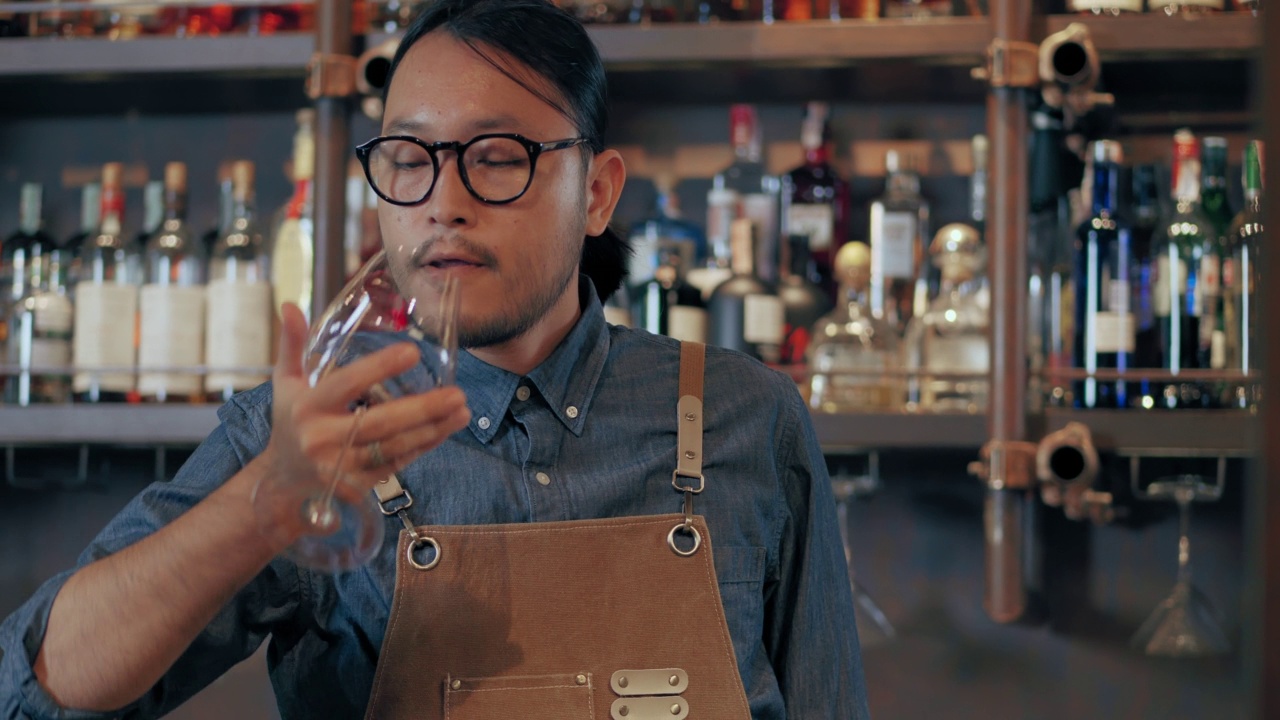 年轻的亚洲侍酒师在品尝新进口的葡萄酒时，微笑着。戴眼镜系领带的男子在酒店的餐厅酒吧品酒。视频下载