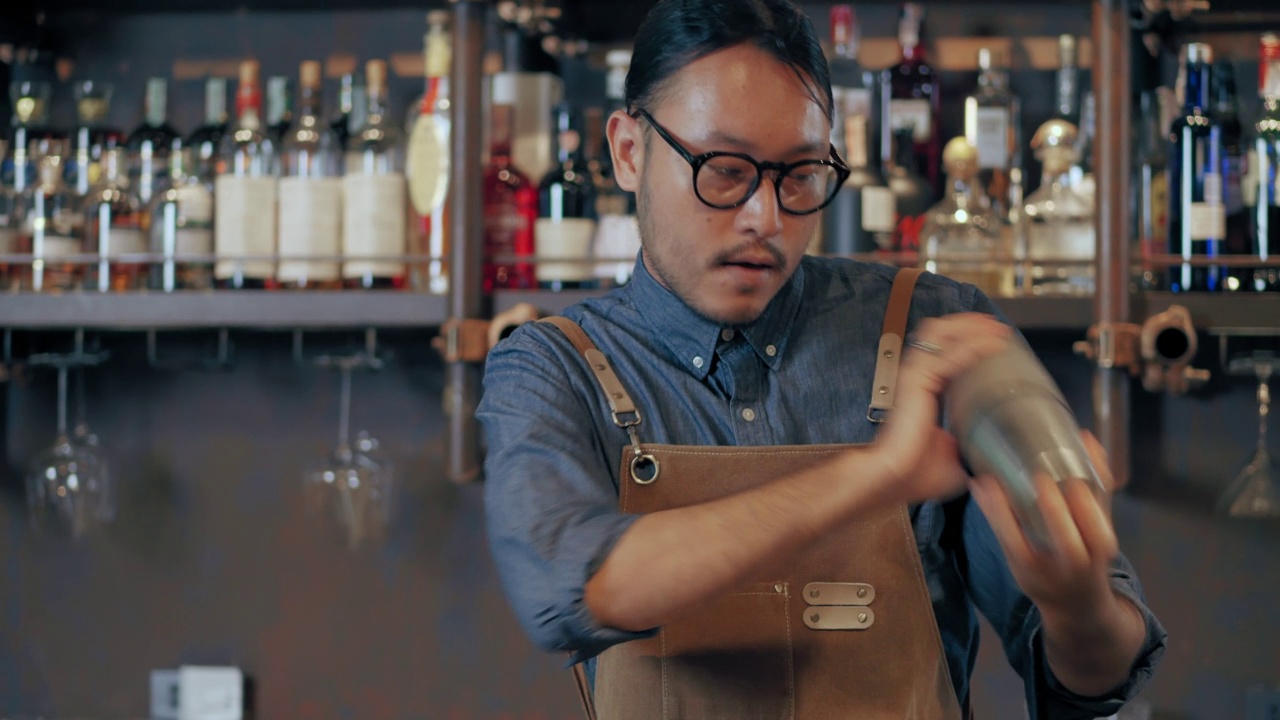 一位年轻的亚洲专业调酒师正在熟练地调制鸡尾酒。一个戴着眼镜、系着围裙的年轻人正在一家酒店餐厅的酒吧里调制鸡尾酒原料。视频下载