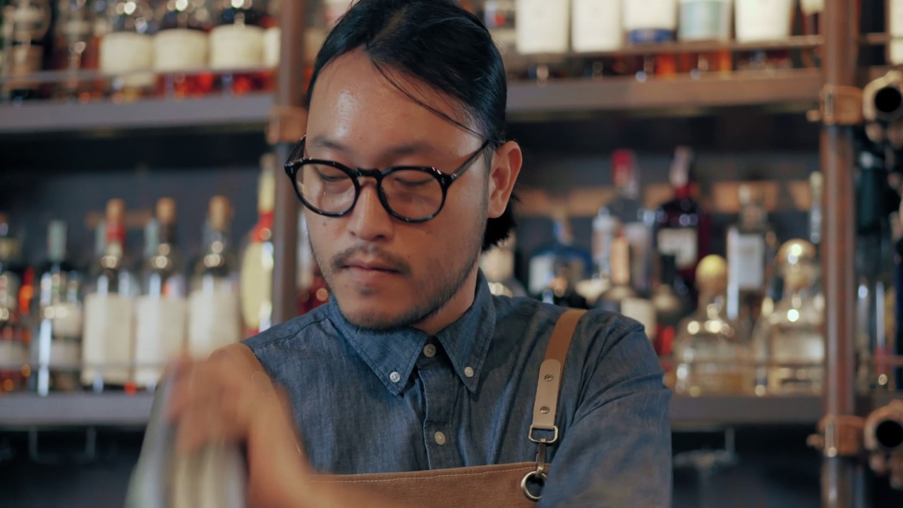 一位年轻的亚洲专业调酒师正在熟练地调制鸡尾酒。一个戴着眼镜、系着围裙的年轻人正在一家酒店餐厅的酒吧里调制鸡尾酒原料。视频素材