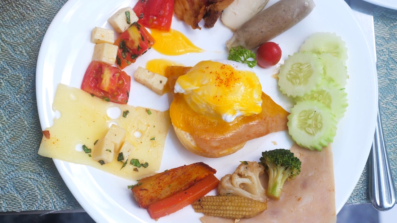 在酒店或度假村的餐厅用手拿着叉子吃早餐。早午餐套餐:荷包蛋、烟熏三文鱼片、荷兰酱、培根、香肠、芝士和烤蔬菜视频下载