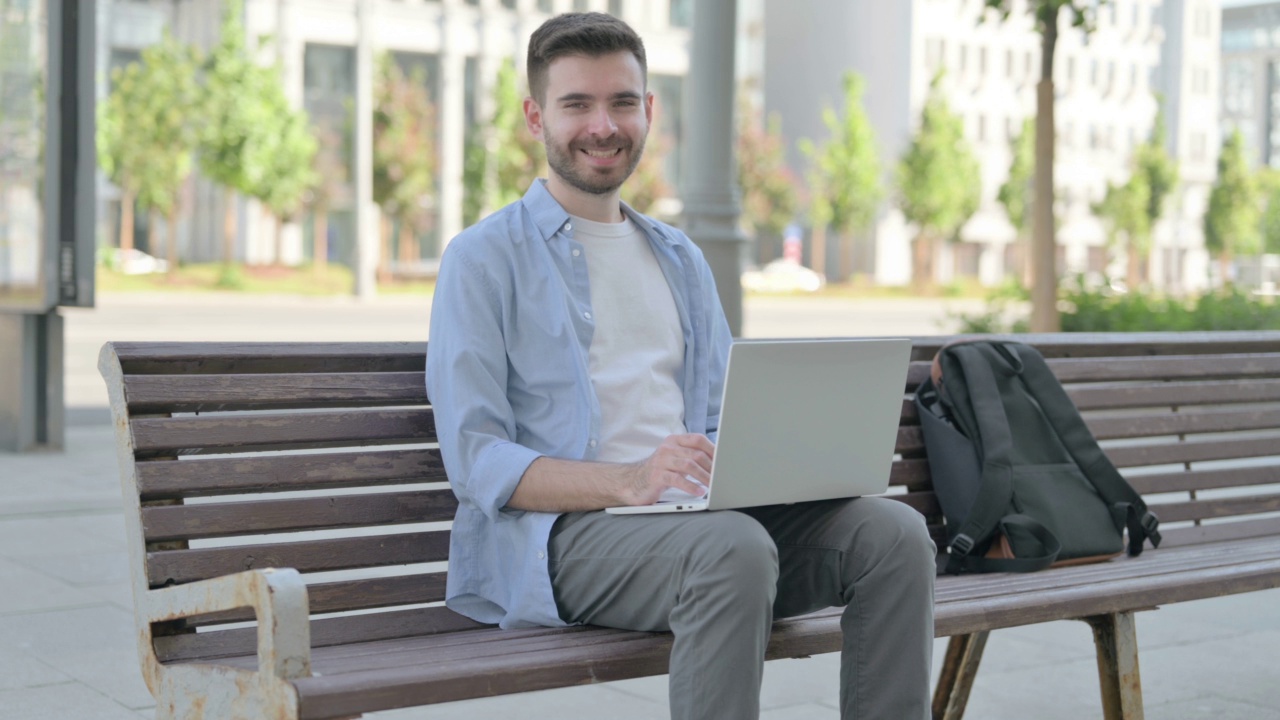 坐在长椅上，带着笔记本电脑的年轻人对着相机微笑视频素材