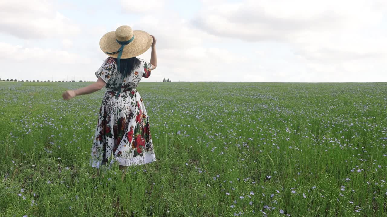 夏天，年轻的女孩戴着大帽子走在开花的亚麻地里。4 k的决议视频下载