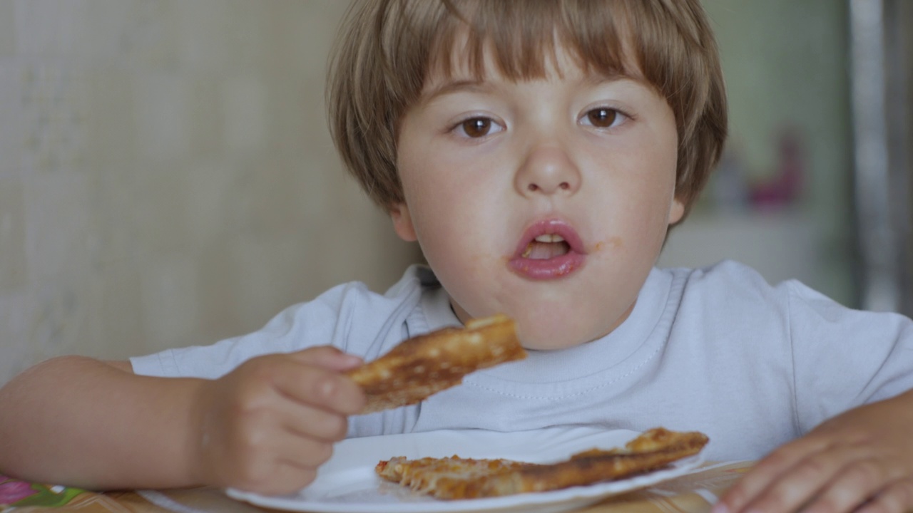 孩子吃披萨。饥肠辘辘的小男孩坐在厨房的餐桌上，吃着盘子里美味的意大利披萨。不健康营养快餐。一个孩子吃午餐的肖像。面对小男孩特写。视频素材