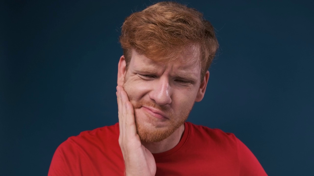 牙痛。英俊的年轻人正在忍受牙痛，用蓝色的背景触摸他的脸颊视频下载