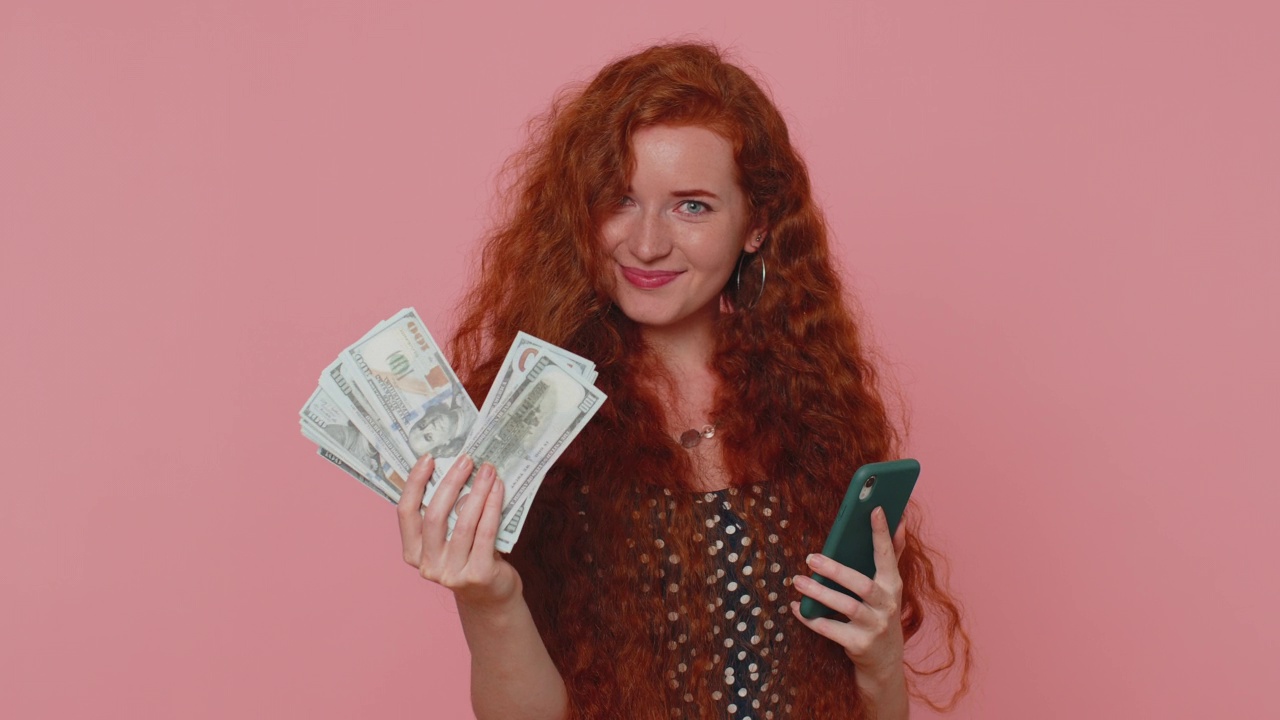 快乐的红发女子看着智能手机显示真诚的喜悦胜利，收到钱美元现金视频素材