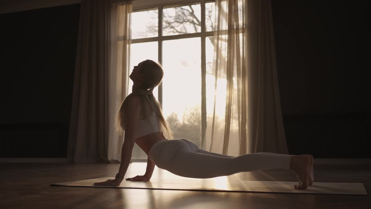 镜头光晕:一名身穿白色运动服的年轻女子在一个有大窗户的大厅里伸展身体，慢镜头拍摄，阳光透过窗户照射进来。健康的生活方式，健康的早晨视频下载