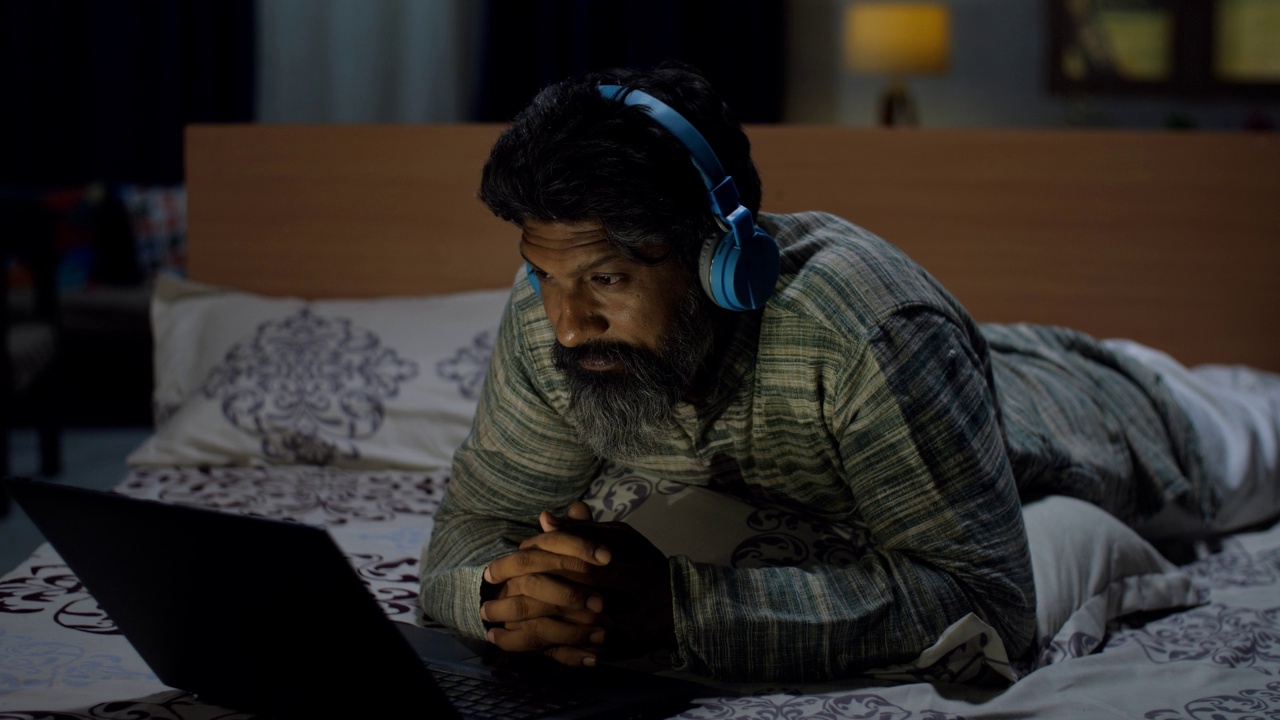 一个留着胡子的印度人戴着耳机在笔记本电脑上看在线视频——深夜视频素材