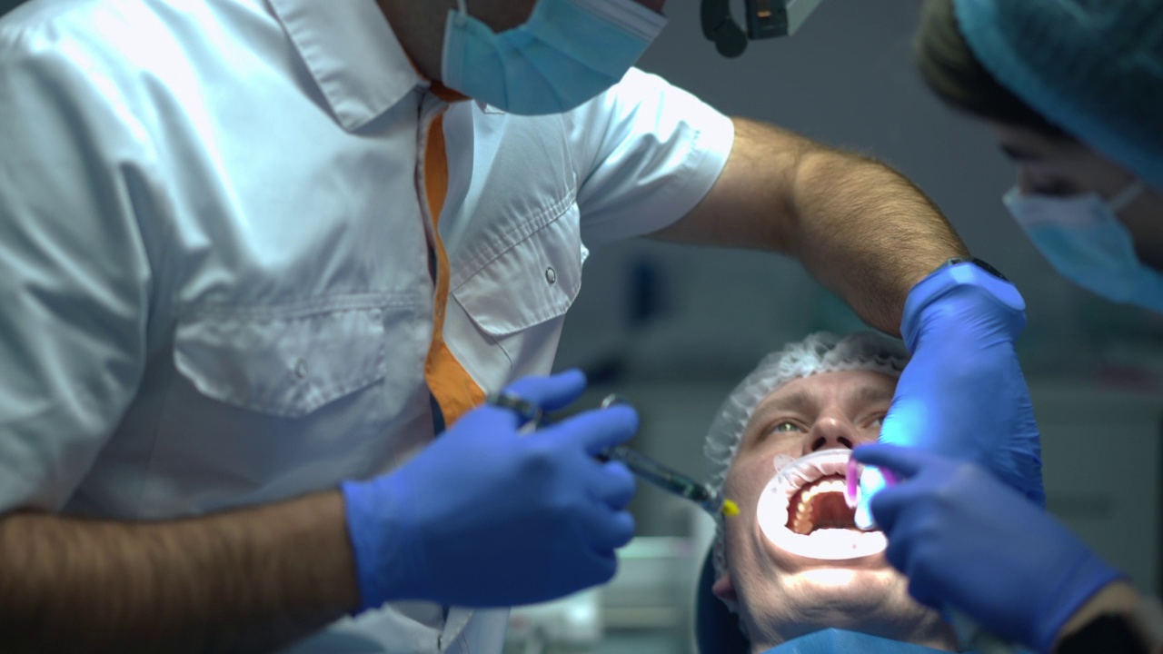 直播摄像机跟踪牙医注射麻醉病人在牙科椅子转动缓慢与注射器。自信的专业医生在助手的帮助下治疗男人。卫生保健。视频素材