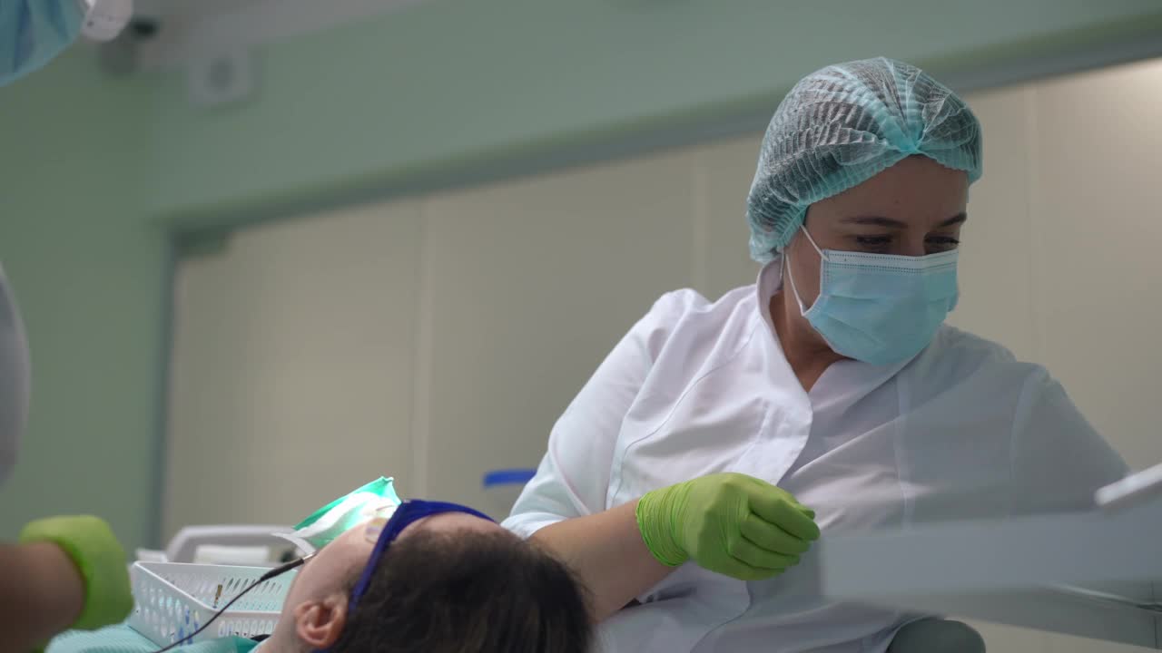 专业牙医戴着口罩治疗病人的慢镜头，助手在前面走过。自信的白人医生和护士在医院与坐在牙科椅上的妇女。视频素材