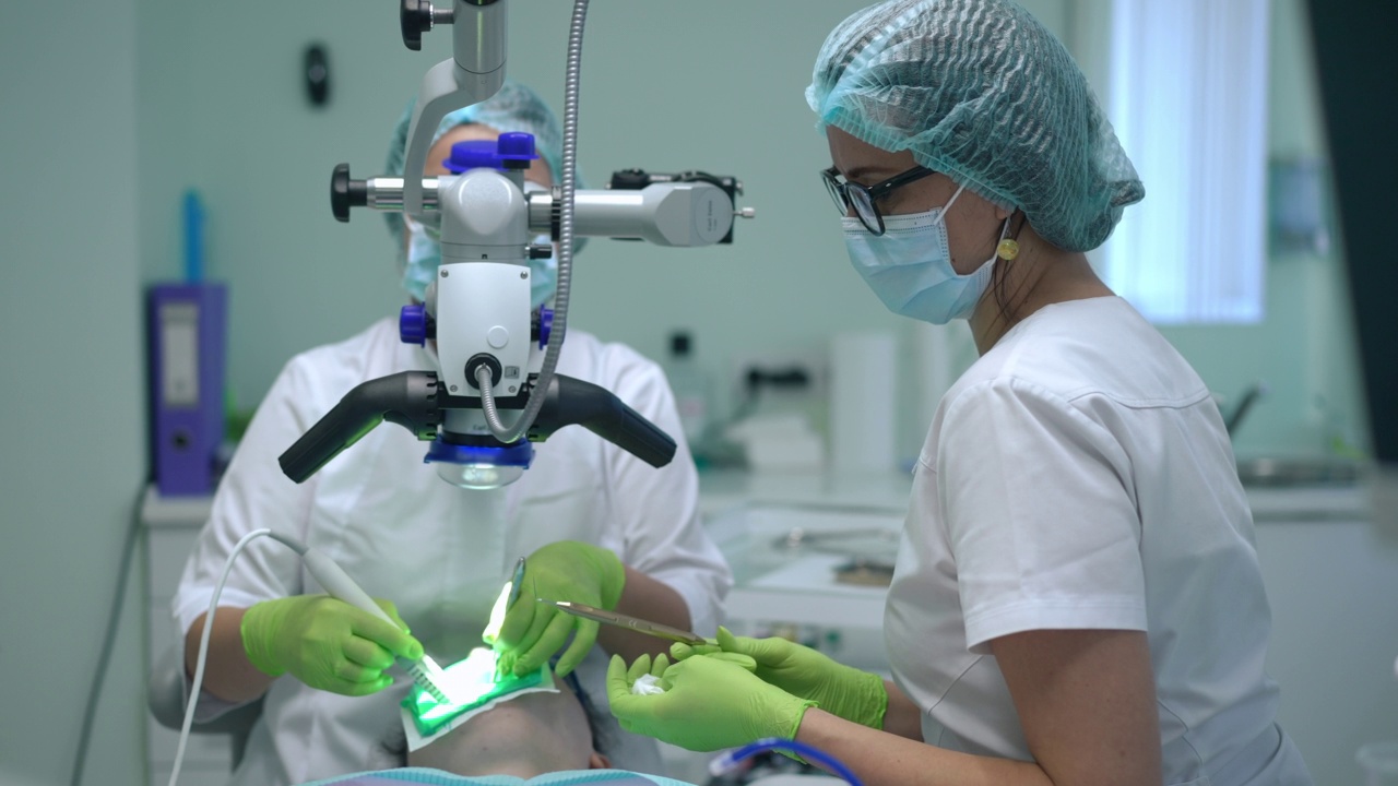 侧视图专业牙科助手在牙科显微镜下给医生递器械的慢镜头。戴口罩的白人妇女在医院协助牙医治疗病人的病牙。视频素材