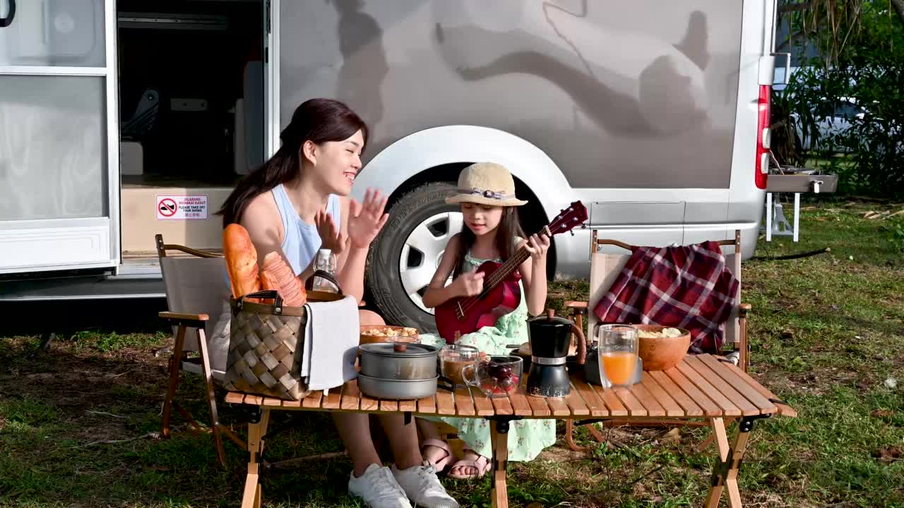 快乐的时刻。一个亚洲小女孩在露营车自驾游的野餐桌上和她的母亲弹吉他视频素材
