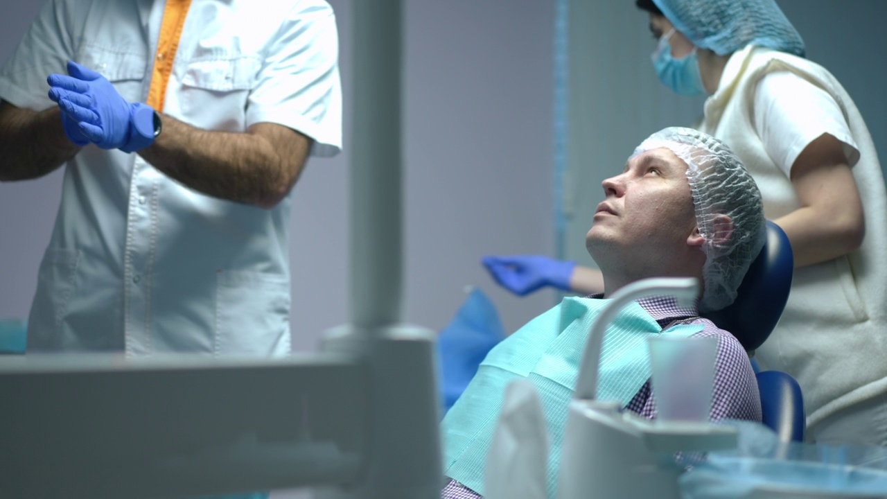 悲伤的白人牙痛男子坐在牙科椅子作为医生护士消毒一次性手套在背景的慢动作。牙齿健康及卫生概念。视频素材