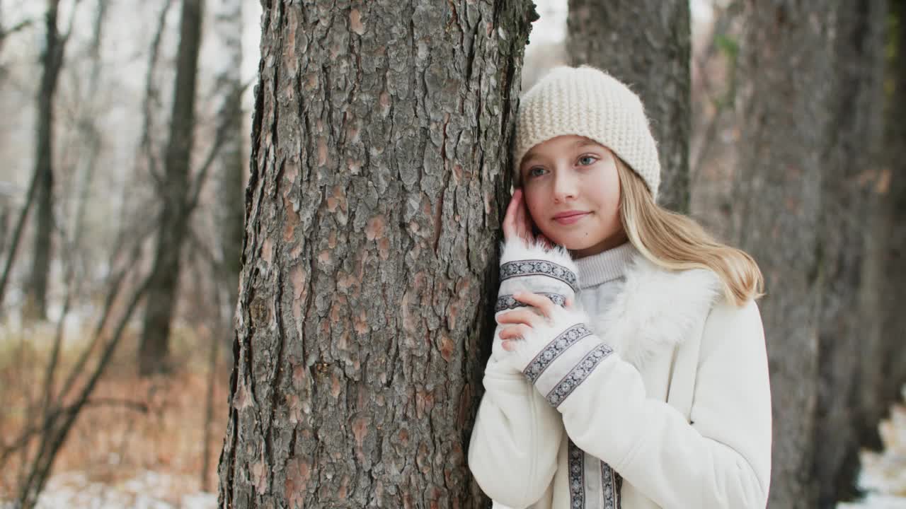 身穿白色夹克、头戴针织帽的浪漫女子倚在树干上拍摄。肖像时尚的女孩在冬季的衣服和帽子摆冬季森林。视频素材