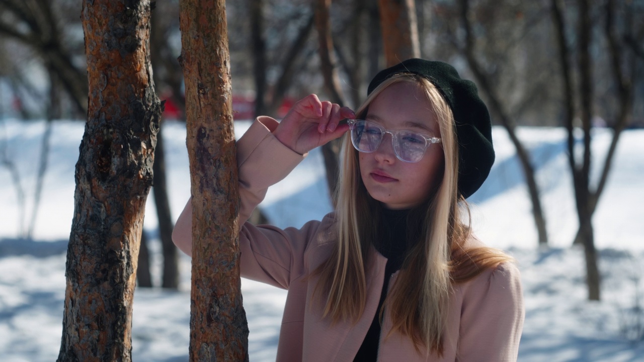 时尚的青少年金发模特女孩在眼镜，粉红色的外套和黑色贝雷帽的背景下模糊的树，雪和移动的汽车在雪地冬季公园附近的树。上半部分。特写镜头。视频下载