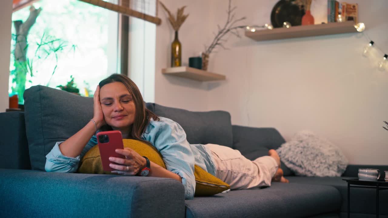 一名年轻的成年女性躺在沙发上使用智能手机视频下载