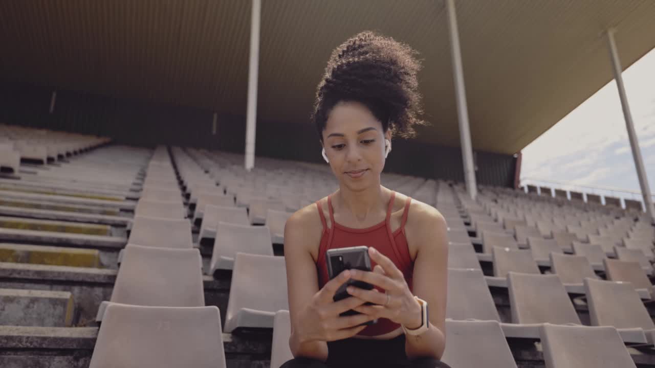 年轻的女运动员在体育馆里用手机发短信，用无线耳机听音乐。运动女性在运动场上训练后独自坐在社交媒体上浏览视频素材