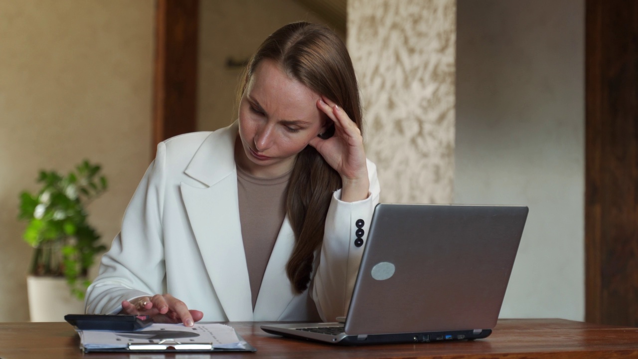 一个孤独的女人穿着白色夹克，背负着很多债务，承受着压力，数着财务，坐在桌子前，拿着纸，笔记本电脑，在计算器上做着计算，试图让收支平衡。视频素材