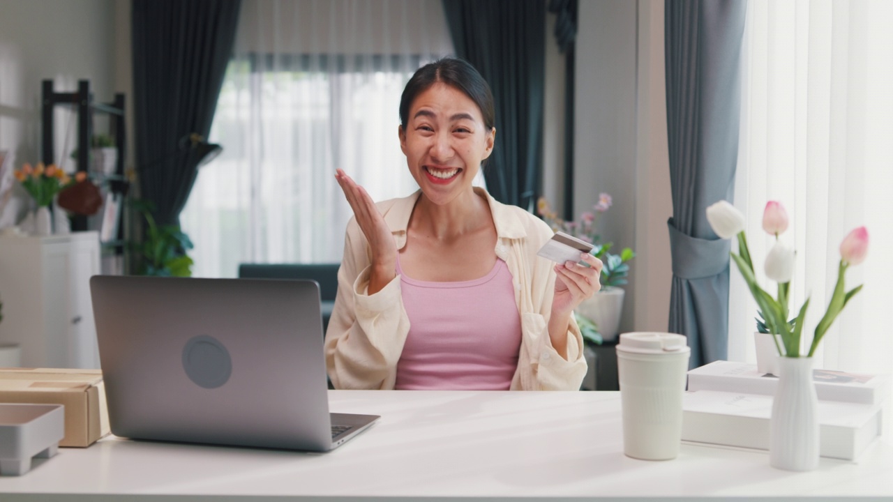 青年亚洲女女孩穿着休闲坐在书桌前与电脑笔记本电脑持有信用卡感到高兴和兴奋的成功付款在网上购物在客厅在家里。视频素材