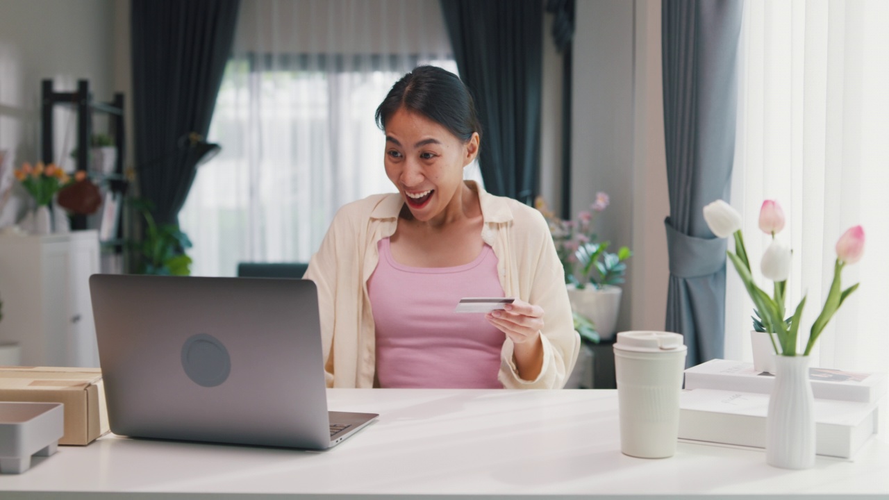 青年亚洲女女孩穿着休闲坐在书桌前与电脑笔记本电脑持有信用卡感到高兴和兴奋的成功付款在网上购物在客厅在家里。视频素材