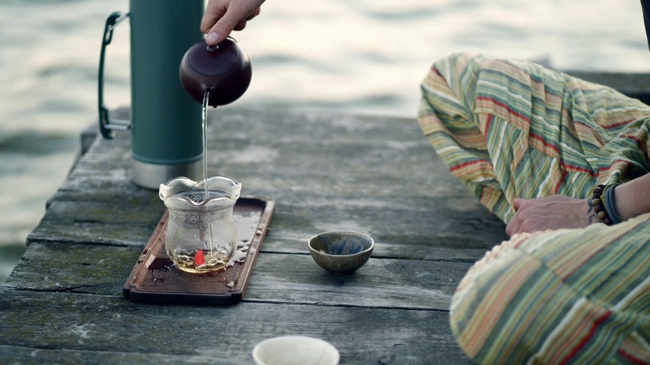 茶道师傅倒青茶。日落时分，岸边茶盘上的茶。4K慢动作视频。以水为背景的绿色保温杯。视频素材