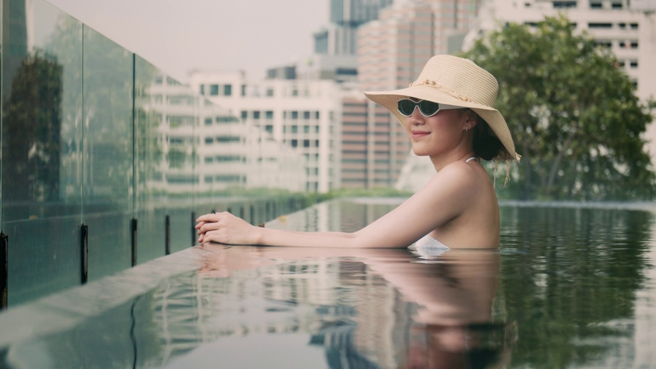 微笑的亚洲女性戴着帽子和太阳镜呆在酒店的游泳池里。视频素材