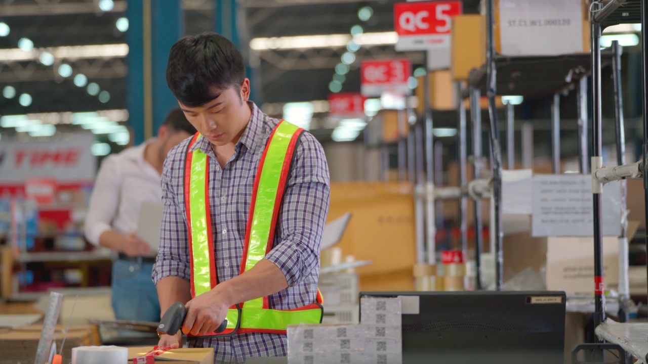 亚洲籍仓库工人在生产线的柜台上进行产品包装，然后使用电脑设置系统，其他白人工人在后面一起工作视频素材