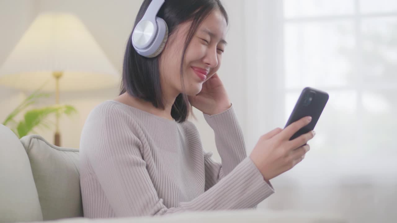 亚洲年轻女性会放松下来，享受她们一起听的音乐。女人度假时戴着耳机坐在沙发上，在家听自己喜欢的歌视频下载