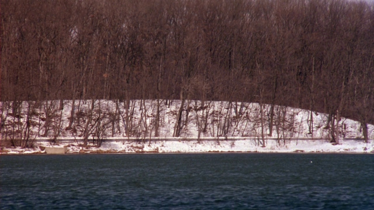 中等角度的湖泊或河流与雪和裸露的树木在岸上。在前景较低处看水，在河岸边看路。查看1930年代的黑色林肯经典汽车从右向左行驶在道路上视频素材