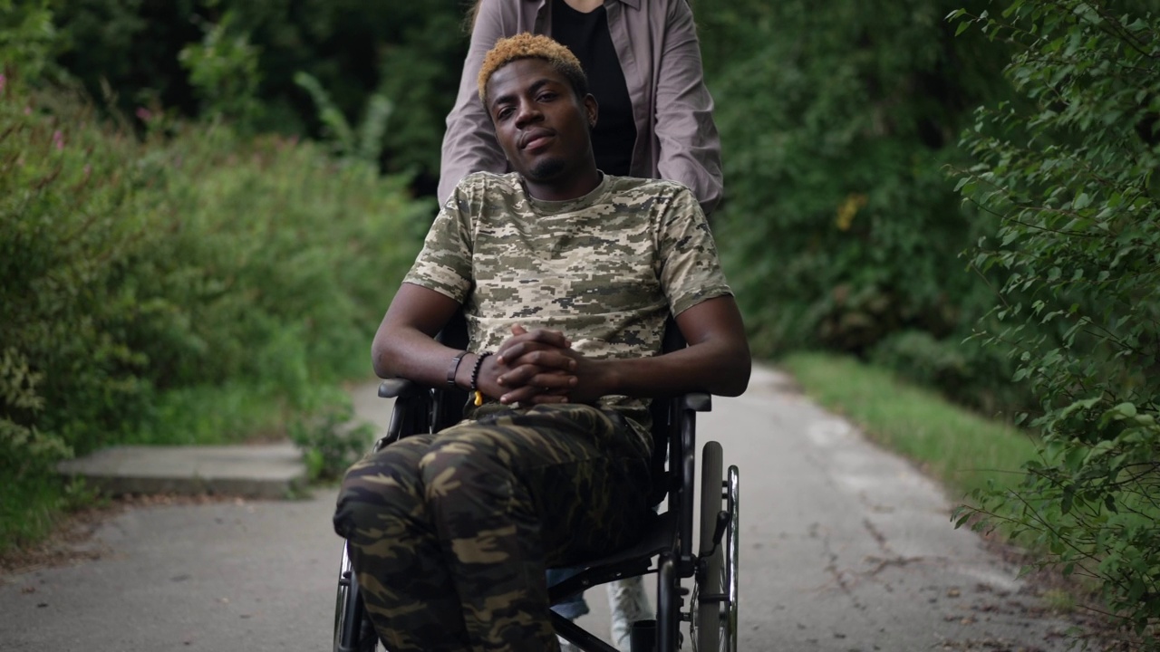 前视图自信的非洲裔美国人在轮椅上与白人妇女骑机动援助设备帮助。多莉拍摄的肖像积极的男朋友在公园和关心的女友。视频素材