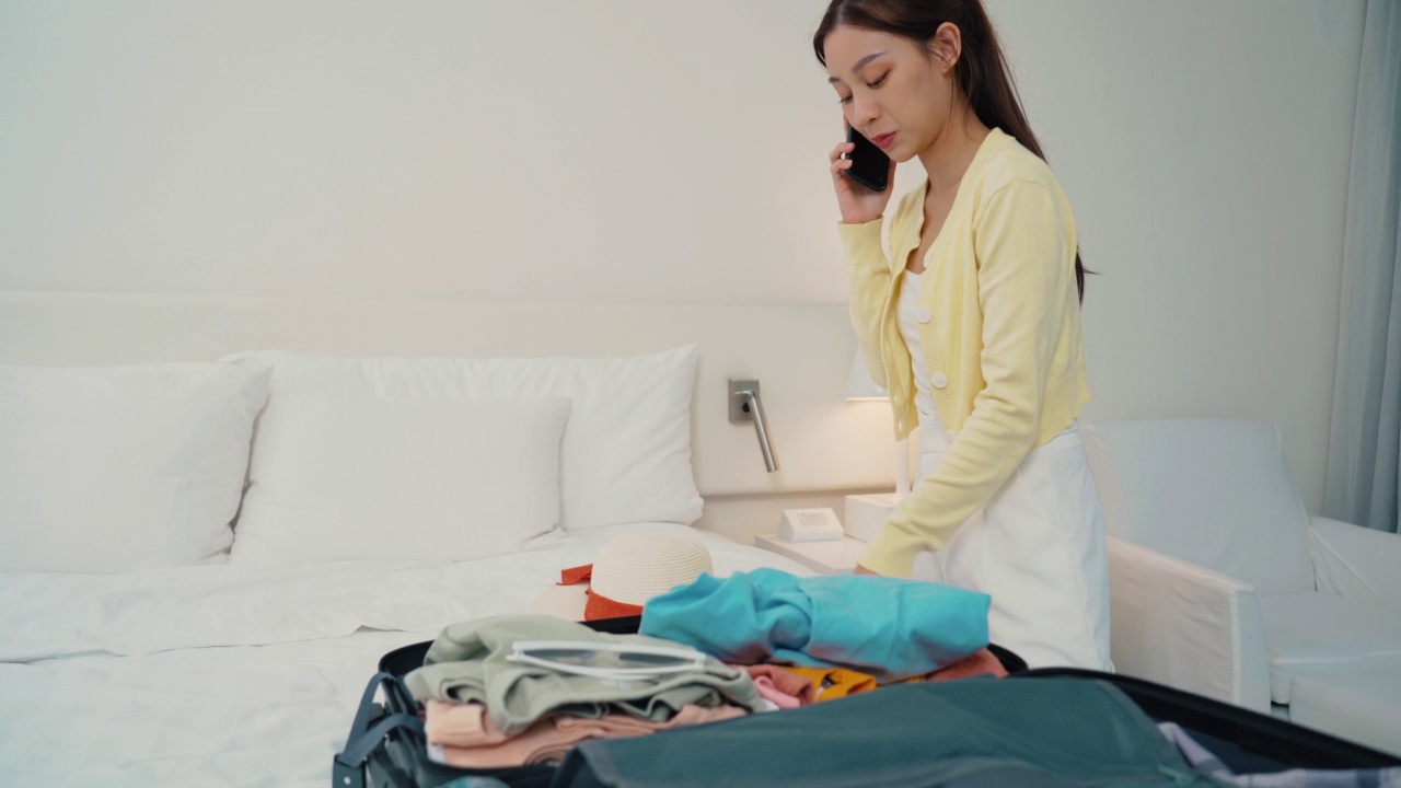 一名亚洲女性把衣服装进行李，在酒店用智能手机打电话视频素材